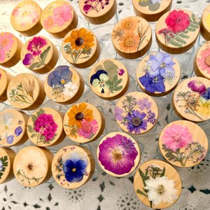 May include: A collection of circular wooden lids for glass jars, each featuring a unique arrangement of pressed dried flowers in vibrant colors including pink, yellow, orange, blue, and purple, displayed in a top-down view.