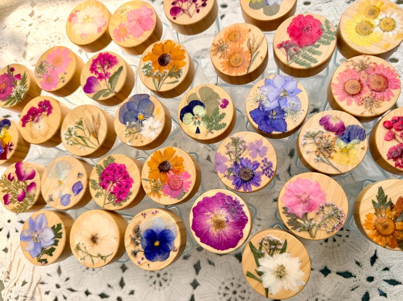 May include: A collection of circular wooden lids for glass jars, each featuring a unique arrangement of pressed dried flowers in vibrant colors including pink, yellow, orange, blue, and purple, displayed in a top-down view.