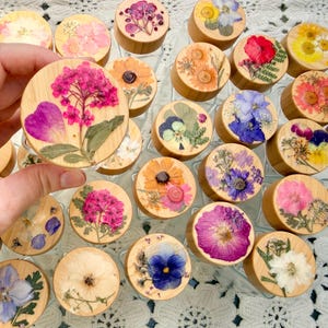 May include: A collection of round wooden jar lids, each featuring a unique arrangement of pressed dried flowers in various colors including pink, purple, yellow, and white. The lids are circular with a smooth, light-toned wood grain finish.