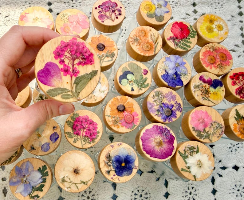 May include: A collection of round wooden jar lids, each featuring a unique arrangement of pressed dried flowers in various colors including pink, purple, yellow, and white. The lids are circular with a smooth, light-toned wood grain finish.
