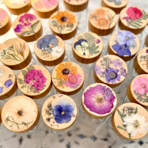May include: A collection of small, round, light-colored wooden lids, each featuring a unique arrangement of pressed, dried flowers in various colors including pink, purple, yellow, blue, and white, set against a clear glass container base.
