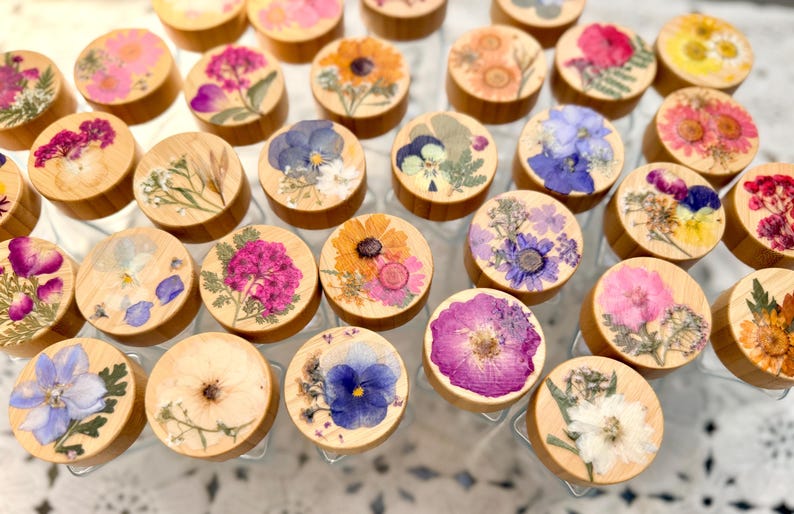 May include: A collection of small, round, light-colored wooden lids, each featuring a unique arrangement of pressed, dried flowers in various colors including pink, purple, yellow, blue, and white, set against a clear glass container base.