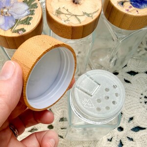 May include: A set of clear glass spice jars with square bases and bamboo lids. Each lid features a unique pressed flower design under a clear resin coating. One jar is shown with its lid removed, revealing a white plastic shaker insert.