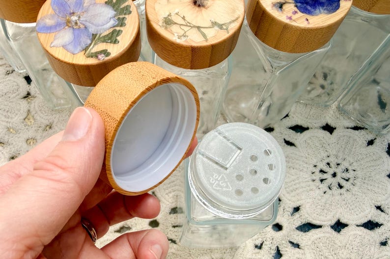May include: A set of clear glass spice jars with square bases and bamboo lids. Each lid features a unique pressed flower design under a clear resin coating. One jar is shown with its lid removed, revealing a white plastic shaker insert.