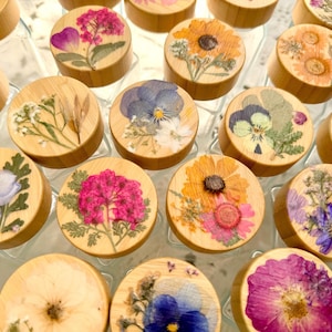 May include: A collection of clear glass jars with round bamboo lids, each featuring a unique arrangement of pressed dried flowers in vibrant colors including pink, yellow, blue, and purple, displayed in a grid pattern from a top-down perspective.