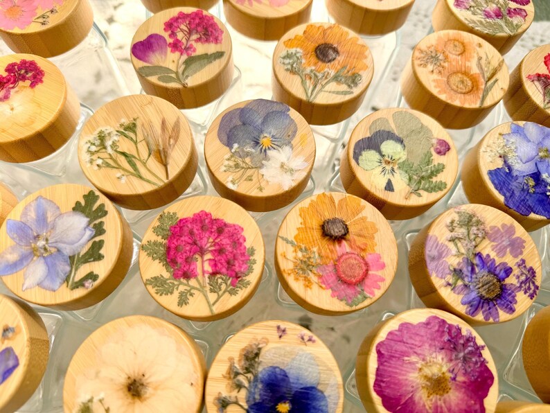 May include: A collection of clear glass jars with round bamboo lids, each featuring a unique arrangement of pressed dried flowers in vibrant colors including pink, yellow, blue, and purple, displayed in a grid pattern from a top-down perspective.