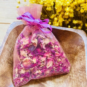 Può includere: Un piccolo sacchetto di rete rosa pieno di petali di rosa secchi, legato con un nastro viola. Il sacchetto è su una superficie di legno.