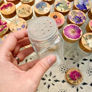 May include: A clear glass spice jar with a square base and a translucent plastic shaker lid. The wooden lid topper is decorated with a pressed flower arrangement featuring various colorful petals and green foliage in shades of pink, blue, and yellow.