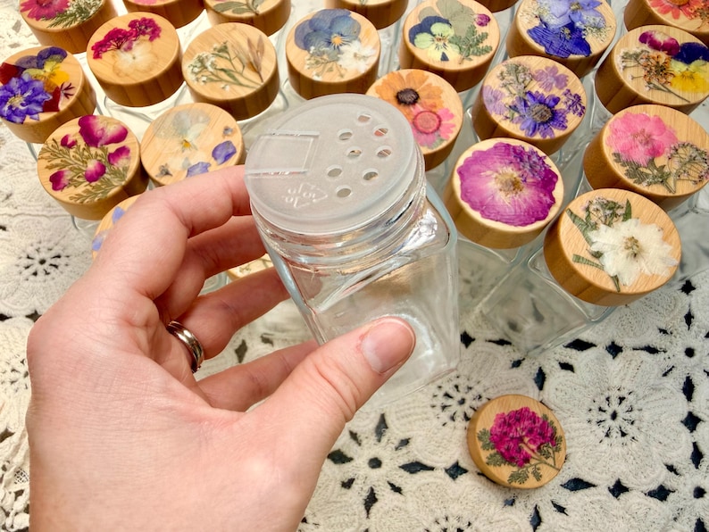 May include: A clear glass spice jar with a square base and a translucent plastic shaker lid. The wooden lid topper is decorated with a pressed flower arrangement featuring various colorful petals and green foliage in shades of pink, blue, and yellow.