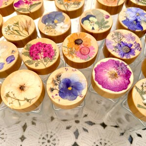 May include: A collection of clear glass jars with round bamboo lids, each featuring a unique arrangement of pressed dried flowers in vibrant shades of pink, blue, yellow, and white, displayed in a grid pattern on a white lace surface.