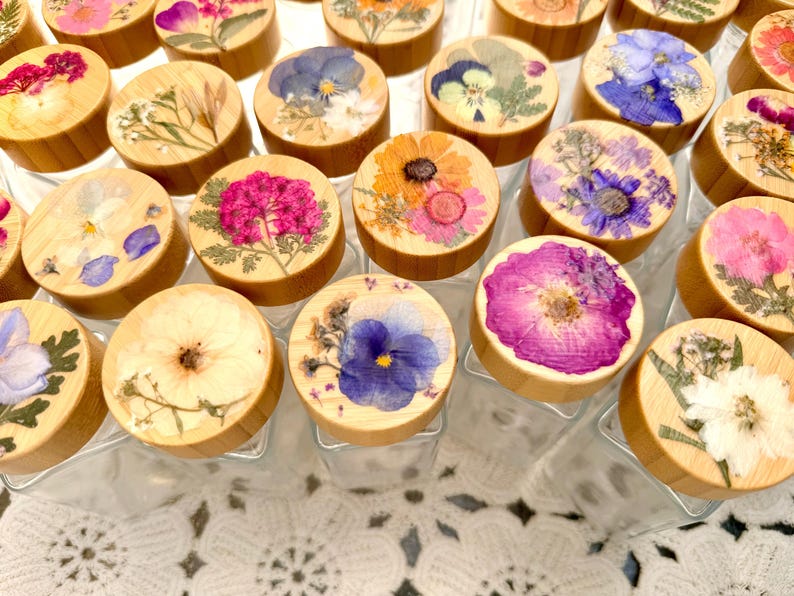 May include: A collection of clear glass jars with round bamboo lids, each featuring a unique arrangement of pressed dried flowers in vibrant shades of pink, blue, yellow, and white, displayed in a grid pattern on a white lace surface.