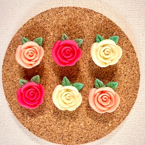 May include: Six colorful rose-shaped embellishments in shades of pink, red, and cream, each with green leaves. The roses are arranged on a round, brown cork surface, creating a decorative floral arrangement.