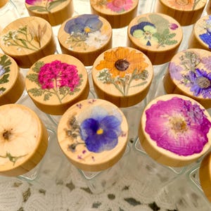May include: A collection of clear glass bottles with round wooden lids, each featuring a unique pressed flower arrangement on top. The flowers display various colors including vibrant pink, deep blue, bright orange, and soft white petals.