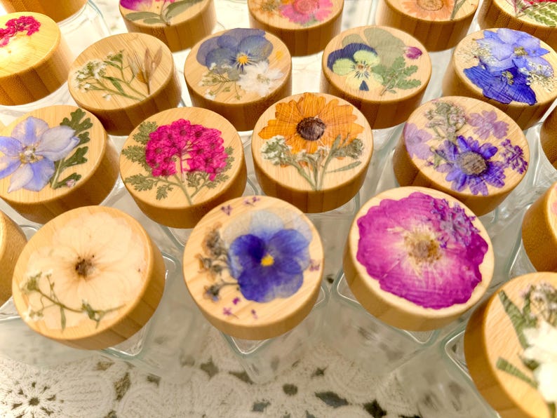 May include: A collection of clear glass bottles with round wooden lids, each featuring a unique pressed flower arrangement on top. The flowers display various colors including vibrant pink, deep blue, bright orange, and soft white petals.