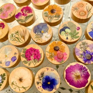 May include: A collection of circular wooden lids, each featuring a unique arrangement of pressed, dried flowers in vibrant shades of pink, yellow, blue, and purple, preserved under a clear, smooth resin finish on the top surface.