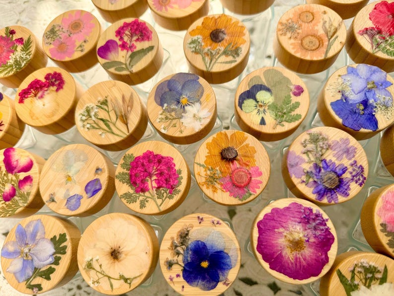 May include: A collection of circular wooden lids, each featuring a unique arrangement of pressed, dried flowers in vibrant shades of pink, yellow, blue, and purple, preserved under a clear, smooth resin finish on the top surface.