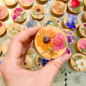 May include: A collection of round wooden jar lids, each featuring a unique arrangement of pressed, dried flowers in vibrant colors including pink, yellow, orange, blue, and white. The lids are smooth, light-toned wood, showcasing natural grain.