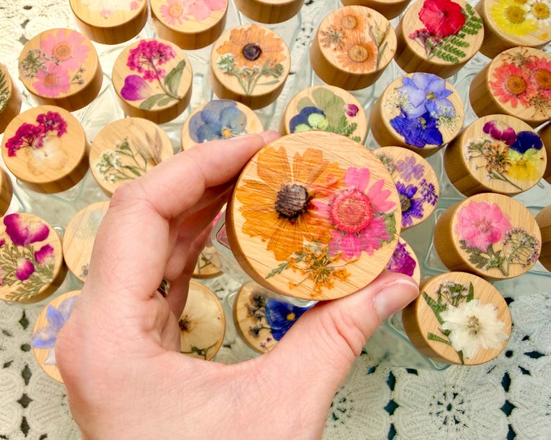 May include: A collection of round wooden jar lids, each featuring a unique arrangement of pressed, dried flowers in vibrant colors including pink, yellow, orange, blue, and white. The lids are smooth, light-toned wood, showcasing natural grain.