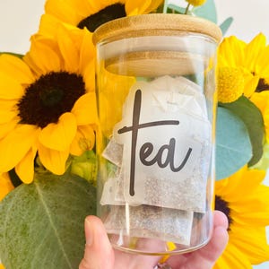 May include: A clear glass jar with a bamboo lid, labeled "Tea" in black script, filled with tea bags. The jar is held in front of a vibrant bouquet of yellow sunflowers and green foliage, creating a bright and cheerful display.
