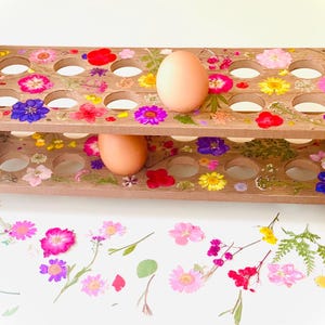 May include: A wooden egg holder with two tiers, decorated with colorful pressed flowers. Two brown eggs rest in the holder, with additional flowers scattered around. The holder has a natural wood finish and a rustic aesthetic.