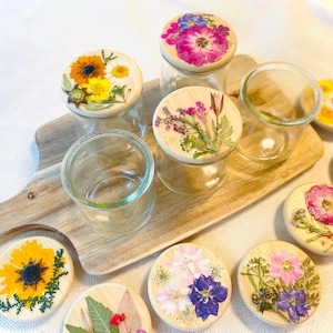 May include: Clear glass jars with wooden lids adorned with pressed flowers. The lids feature various floral arrangements in shades of yellow, pink, purple, and white. The jars sit on a wooden serving board, with additional lids scattered around.