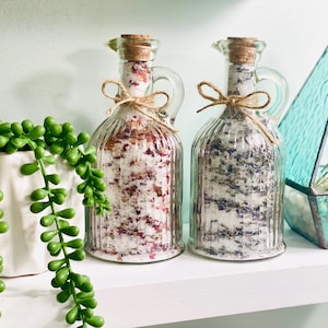 May include: Two clear glass bottles with cork stoppers filled with bath salts. The bottle on the left contains pink bath salts with dried rose petals. The bottle on the right contains lavender bath salts. Both bottles have a rustic twine and burlap bow.