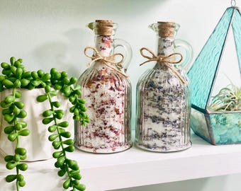 May include: Two clear glass bottles with cork stoppers filled with bath salts. The bottle on the left contains pink bath salts with dried rose petals. The bottle on the right contains lavender bath salts. Both bottles have a rustic twine and burlap bow.