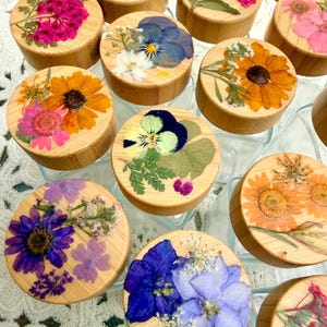 May include: A collection of round bamboo lids for glass jars, each featuring a unique arrangement of pressed dried flowers in vibrant colors including pink, orange, blue, purple, and yellow, set against the natural light wood grain surface.