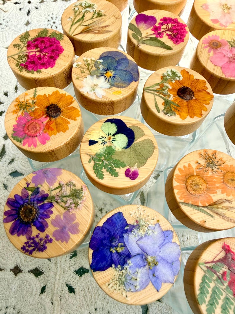 May include: A collection of round bamboo lids for glass jars, each featuring a unique arrangement of pressed dried flowers in vibrant colors including pink, orange, blue, purple, and yellow, set against the natural light wood grain surface.