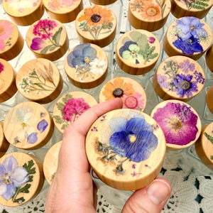 May include: A collection of clear glass jars with round wooden lids, each featuring a unique arrangement of pressed dried flowers in vibrant colors like blue, pink, yellow, and white, preserved under a clear resin coating on the top surface.