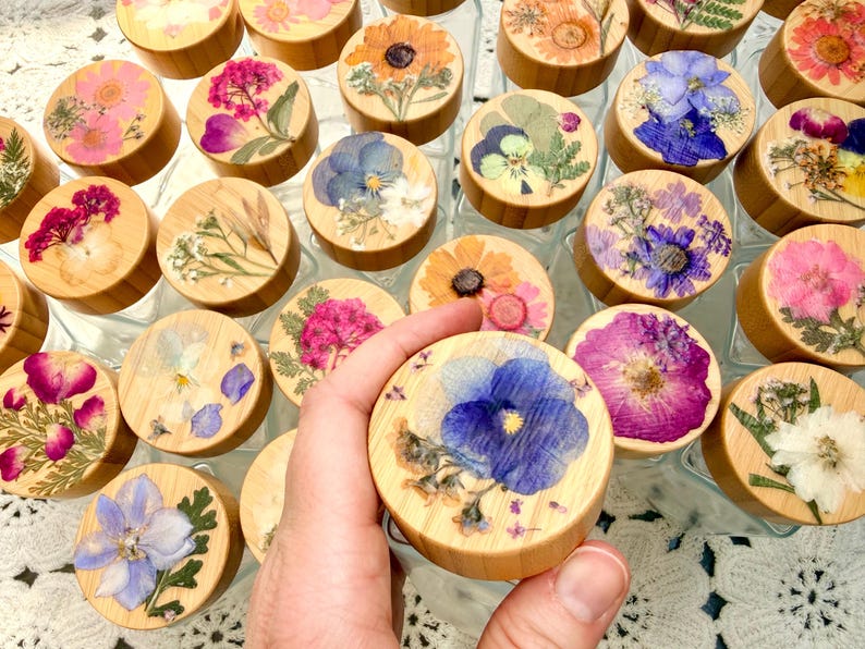May include: A collection of clear glass jars with round wooden lids, each featuring a unique arrangement of pressed dried flowers in vibrant colors like blue, pink, yellow, and white, preserved under a clear resin coating on the top surface.