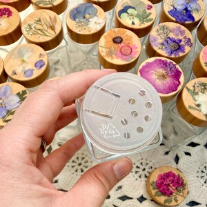 May include: A clear glass spice jar with a square base and a translucent plastic shaker lid featuring a hinged flap and seven circular holes. The jar is surrounded by multiple wooden lids, each decorated with a unique pressed flower arrangement.