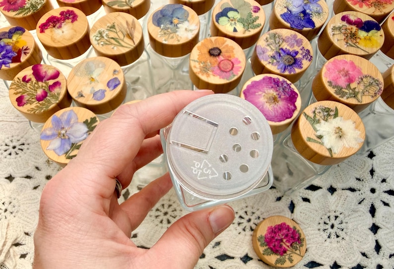 May include: A clear glass spice jar with a square base and a translucent plastic shaker lid featuring a hinged flap and seven circular holes. The jar is surrounded by multiple wooden lids, each decorated with a unique pressed flower arrangement.