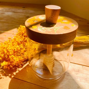 May include: A clear glass honey jar with a wooden lid and dipper. The lid is decorated with yellow floral designs. The jar sits on a wooden surface, with dried yellow flowers in the background.