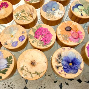 May include: A collection of glass jars with round bamboo lids, each featuring a unique arrangement of pressed dried flowers in vibrant shades of pink, blue, yellow, and white, sealed under a clear resin coating on the top surface.