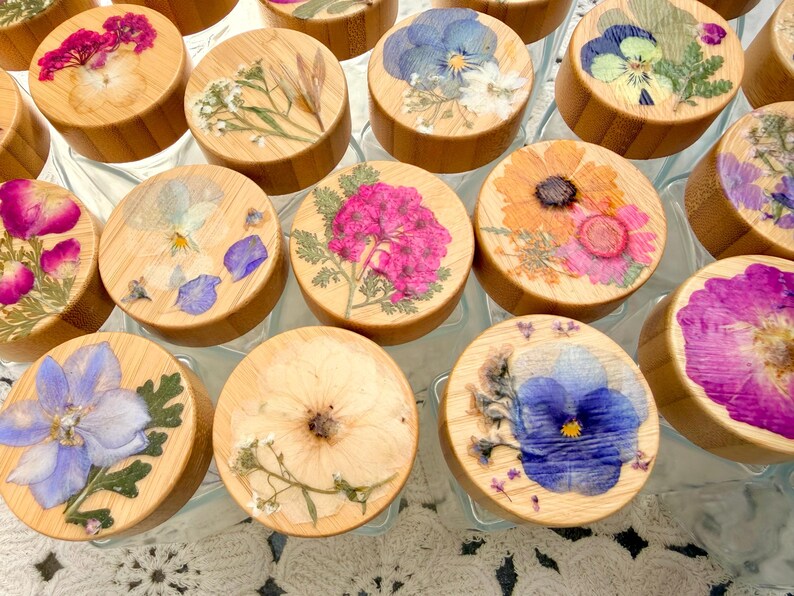 May include: A collection of glass jars with round bamboo lids, each featuring a unique arrangement of pressed dried flowers in vibrant shades of pink, blue, yellow, and white, sealed under a clear resin coating on the top surface.