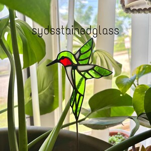 May include: A stained glass hummingbird with green wings, a red throat, and a black beak. The hummingbird is mounted on a metal stake, perfect for a garden or indoor plant. The text "sydsstainedglass" is in the upper left corner.