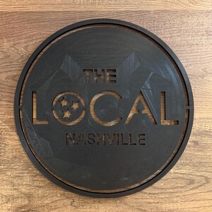 Peut inclure: Une enseigne circulaire noire avec le texte "The Local Nashville" en blanc, découpé. L'enseigne présente un motif d'étoile stylisé.