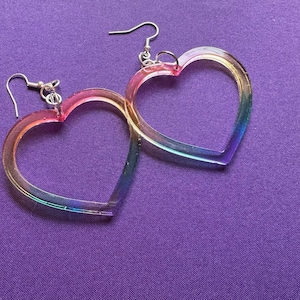 May include: A pair of heart-shaped earrings with a rainbow gradient. The earrings have a clear, iridescent appearance with silver-colored ear wires. The earrings are displayed on a purple background.