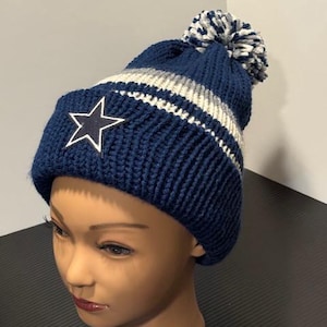 May include: A navy blue knit beanie with a white star emblem and a white outline. The hat features white and gray stripes and a matching pom-pom. The beanie is displayed on a mannequin head.
