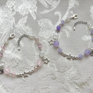 May include: Two silver-toned chain bracelets featuring round beads and star-shaped charms. One bracelet displays light pink beads and clear star beads, while the other features purple beads and clear star beads. Both include a small spiral metal charm.