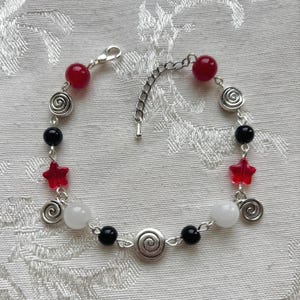 Puede incluir: Una pulsera de cadena en tono plateado con una mezcla de cuentas redondas rojas, negras y blancas, cuentas rojas en forma de estrella y dijes de espiral plateados. La pulsera incluye un cierre de langosta y una cadena extensora ajustable.
