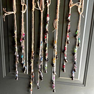 May include: A set of six vertical hanging strands made of brown twine, each featuring multiple small dark metal bells interspersed with colorful translucent beads in shades of red, blue, green, yellow, and purple, suspended against a dark door.