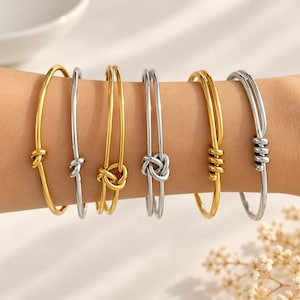 Puede incluir: Una colección de seis brazaletes de metal usados juntos, que incluye tres pulseras en tono dorado y tres en tono plateado. Cada brazalete presenta un detalle de nudo distintivo, que varía desde bucles simples hasta complejos patrones de nudos.