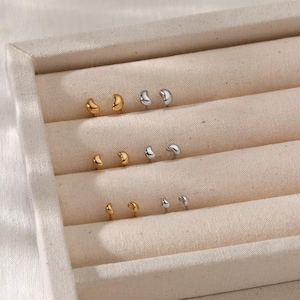 May include: Three pairs of small, smooth, bean-shaped stud earrings arranged in a row. The top and bottom pairs are gold-toned, while the middle pair is silver-toned. Each earring features a curved, polished surface with a simple, rounded design.