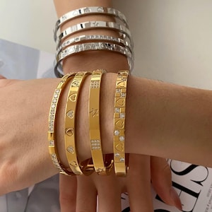 Puede incluir: Una colección de cuatro brazaletes plateados y cuatro dorados en las muñecas. Los brazaletes plateados presentan recortes de corazones y estrellas con piedras claras. Los brazaletes dorados muestran patrones de corazones, estrellas y cuadrados.