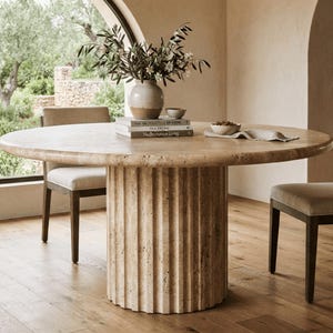 May include: A round beige travertine dining table with a thick, fluted cylindrical pedestal base. The table is accompanied by two chairs with dark wooden frames and light-coloured upholstered seats. A ceramic vase with greenery sits on top.
