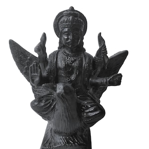 Puede incluir: Una estatua negra de una deidad con cuatro brazos, que lleva una corona y joyas ornamentadas, sentada sobre un pájaro grande con las alas extendidas. La figura tiene una mano levantada en gesto de bendición y está montada sobre una base rectangular.