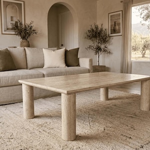 Puede incluir: Una mesa de centro rectangular hecha de piedra travertino beige con una superficie superior lisa y plana y cuatro patas cilíndricas gruesas. La mesa presenta una textura porosa natural y variaciones de color tostado claro.
