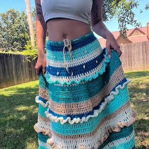 Puede incluir: Una falda larga de crochet de varias capas con un patrón de rayas azules, blancas y marrones. La falda tiene un dobladillo con volantes y está decorada con pequeñas conchas.